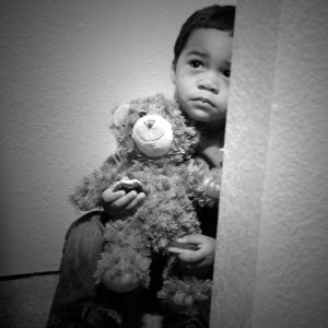 An unhappy-looking little boy sits with his teddy bear on the floor of a closet.