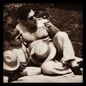 A father and his young son sit together on a blanket on the lawn on a sunny day. Each have their shirts removed and are dressed almost identically including straw hats, sunglasses, and pipes." title="A father and his young son sit together on a blanket on the lawn on a sunny day. Each have their shirts removed and are dressed almost identically including straw hats, sunglasses, and pipes.