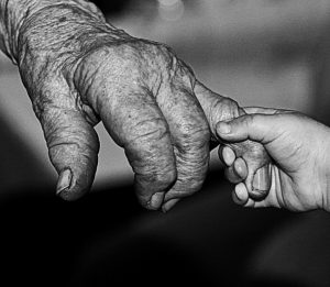 A very small hand of a child holds a wrinkled finger on the hand of a very old person.
