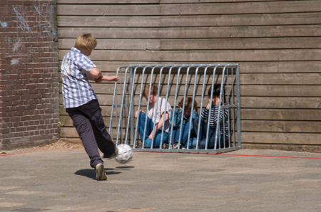 Young kids hide behind a hockey net while another boy kicks a ball at them.