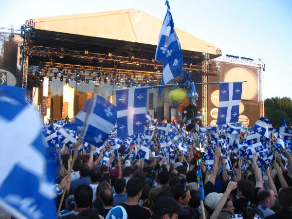 A crowd of people stand in front of a stage. Many hold Quebec flags in the air.