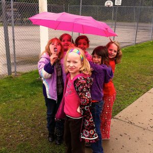 A group of young girls stand together under an umbrella on the playground.