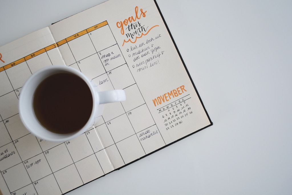 A photo of a coffee cup sitting on a calendar.  The calendar has a section for goals this month.